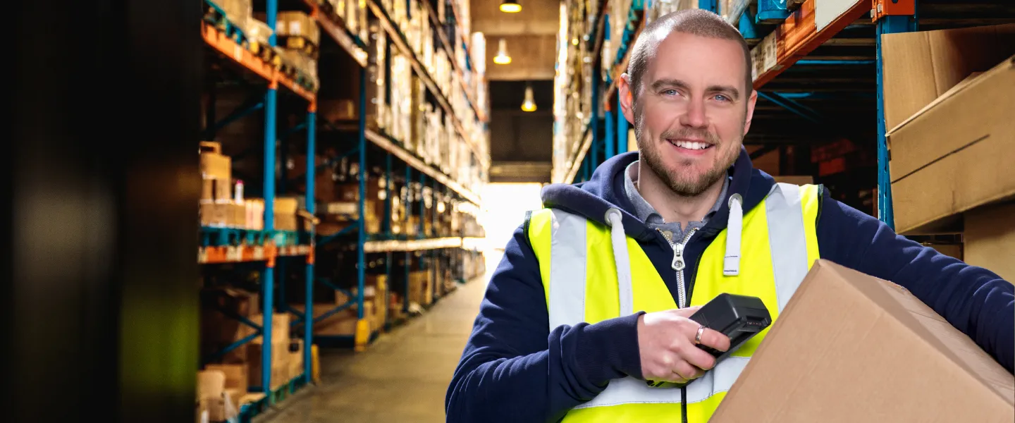 Pracownik magazynu w kamizelce odblaskowej trzymający skaner i karton, stojący między regałami z towarem.