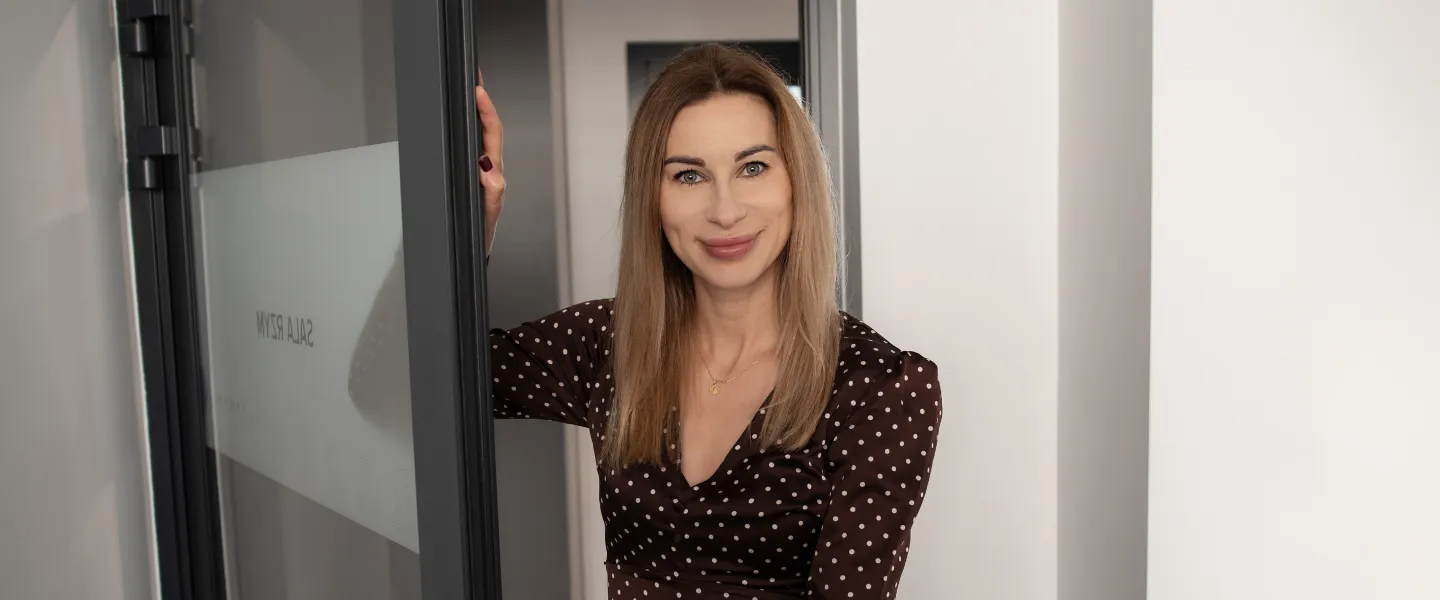 Kobieta stojąca w otwartych drzwiach sali konferencyjnej, uśmiechnięta i patrząca w stronę aparatu.