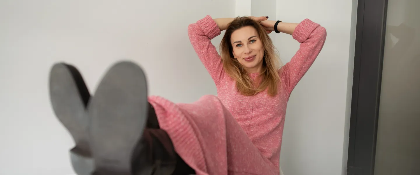 Uśmiechnięta kobieta w różowej dzianinowej sukience siedzi w jasnym wnętrzu, z rękami za głową i nogami wyciągniętymi w stronę obiektywu.