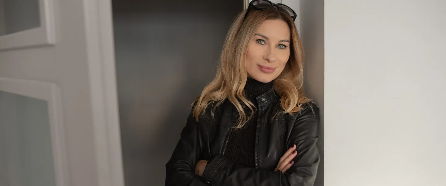 Kobieta o pewnej postawie, ubrana w czarną skórzaną kurtkę, stoi oparta o ścianę; patrzy z lekkim uśmiechem, reprezentując autentyczne przywództwo, odwagę i przełamywanie schematów