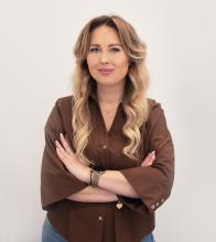 Kobieta o falowanych blond włosach w brązowej koszuli stoi na białym tle, z rękami skrzyżowanymi, lekko się uśmiecha.